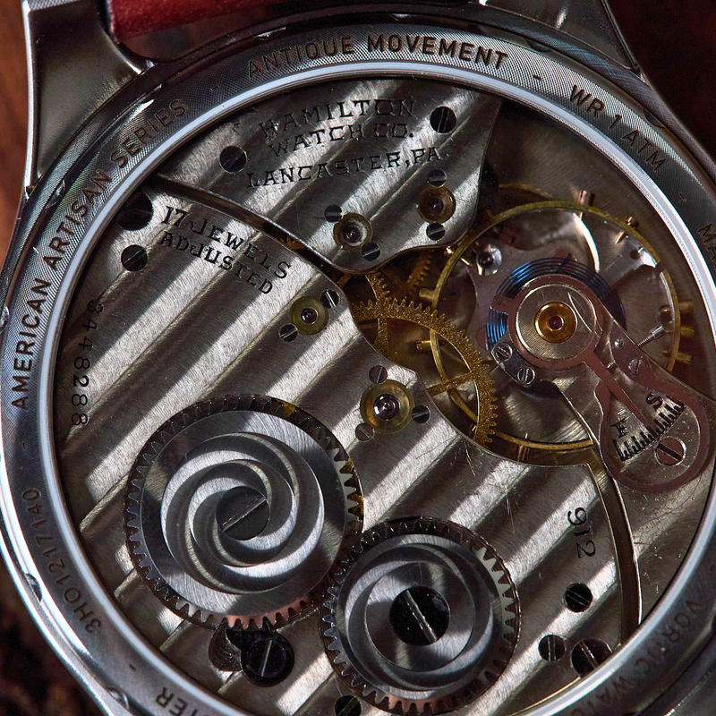 Close-up of a watch movement showing the silver-toned case back, internal gears, bridges, and hands, with engraved text including 'AMERICAN ARTISAN SERIES' and 'ANTIQUE MOVEMENT'.