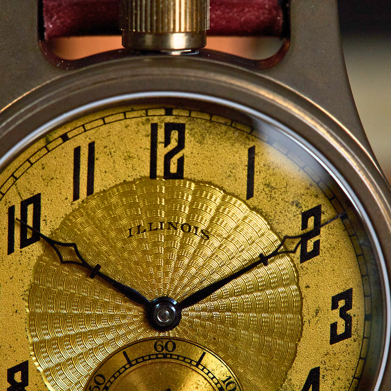 Close-up of a vintage-style watch with a round metallic case, black numerals, and a textured gold dial. The brand name 'ILLINOIS' is visible on the dial.