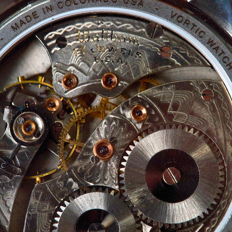 Close-up of a vintage watch movement with engraved steel components, gears, and copper-colored screws, showing text including 'THE BOSTON' and 'VORTEX WATCH'.