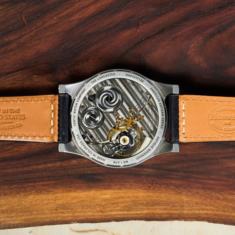 A close-up of a wristwatch laid flat on a dark wood surface, showing the open back with a visible mechanical movement and a tan leather strap with black edges.