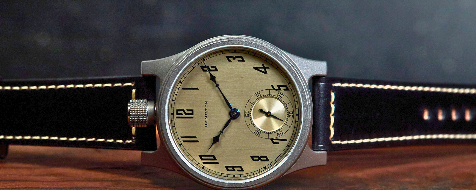 A vintage-style wristwatch with a round silver-toned case, beige dial, black leather strap with white contrast stitching, and a small gold-tone subdial, resting on a wooden surface against a dark textured background.