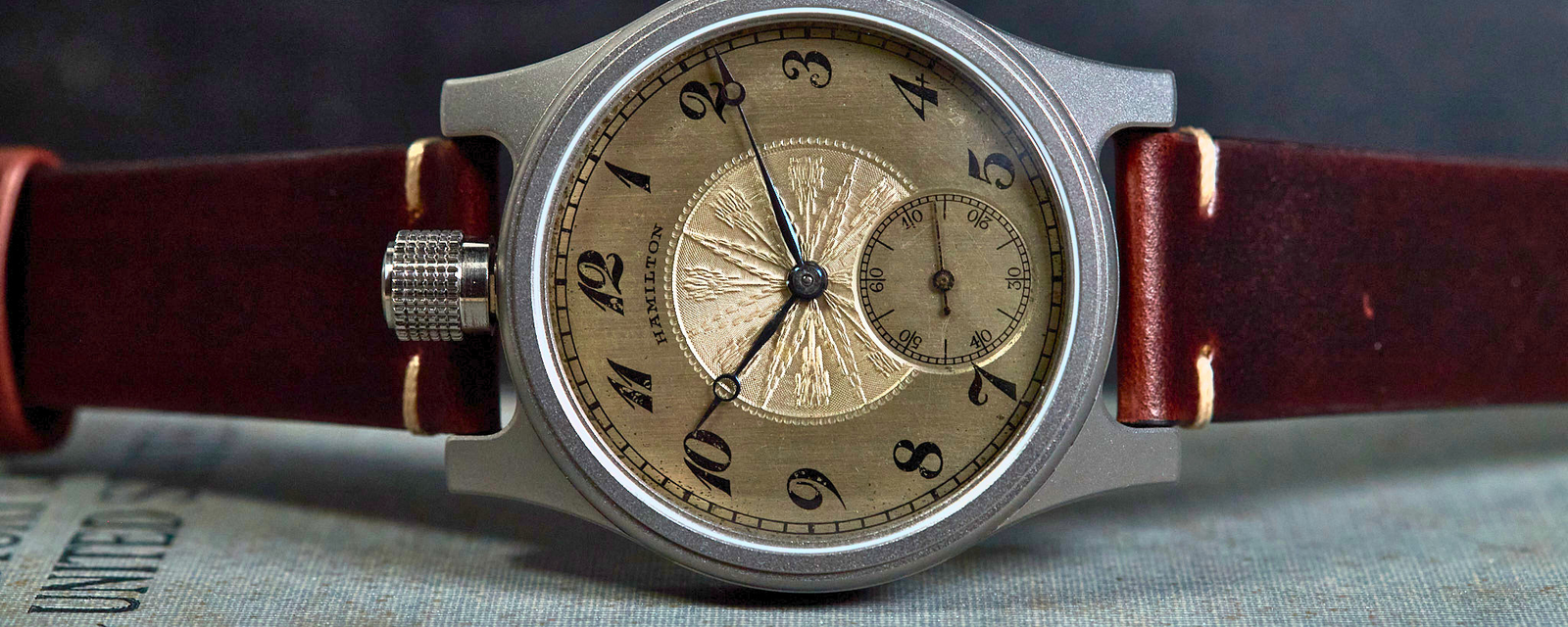 A vintage-style wristwatch with a silver-toned metal case, a beige sunburst dial with black Arabic numerals, and a dark reddish-brown leather strap, resting on a textured blue-gray surface against a dark background.