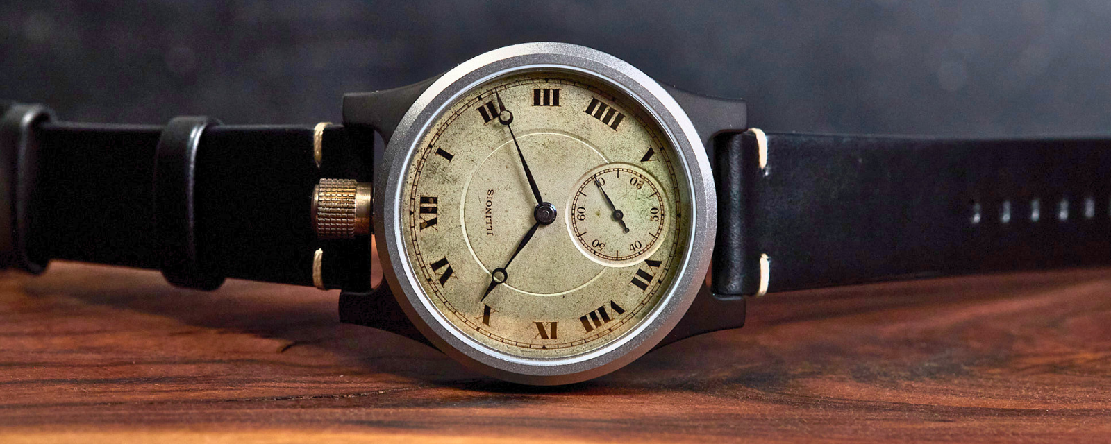 A vintage-style wristwatch with a round silver-tone case, cream dial with black Roman numerals, and a black leather strap, resting on a rustic wooden surface against a dark blurred background.