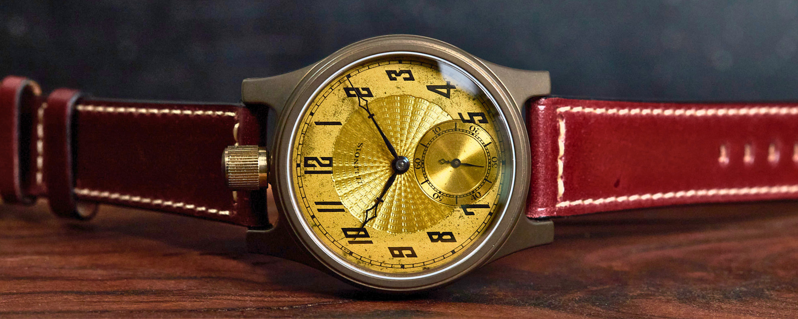 A wristwatch with a round gold sunburst dial, black Arabic numerals, and a brushed metal case, shown resting on a wooden surface with a dark textured background. The watch has a reddish-brown leather strap with white contrast stitching.