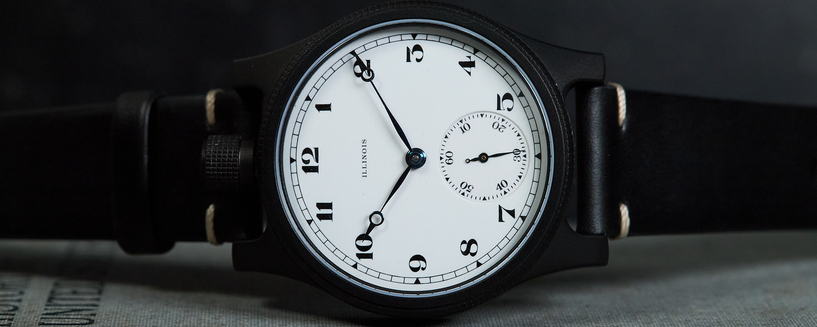 A black dial watch with a white face and black strap, resting on a textured gray surface against a dark background.
