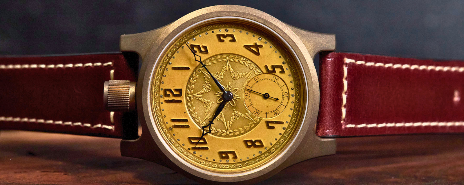 A vintage-style wristwatch with a round gold-toned dial, black Arabic numerals, a textured central subdial, a brushed metal case, and a reddish-brown leather strap with white stitching, resting on a wooden surface against a dark background.