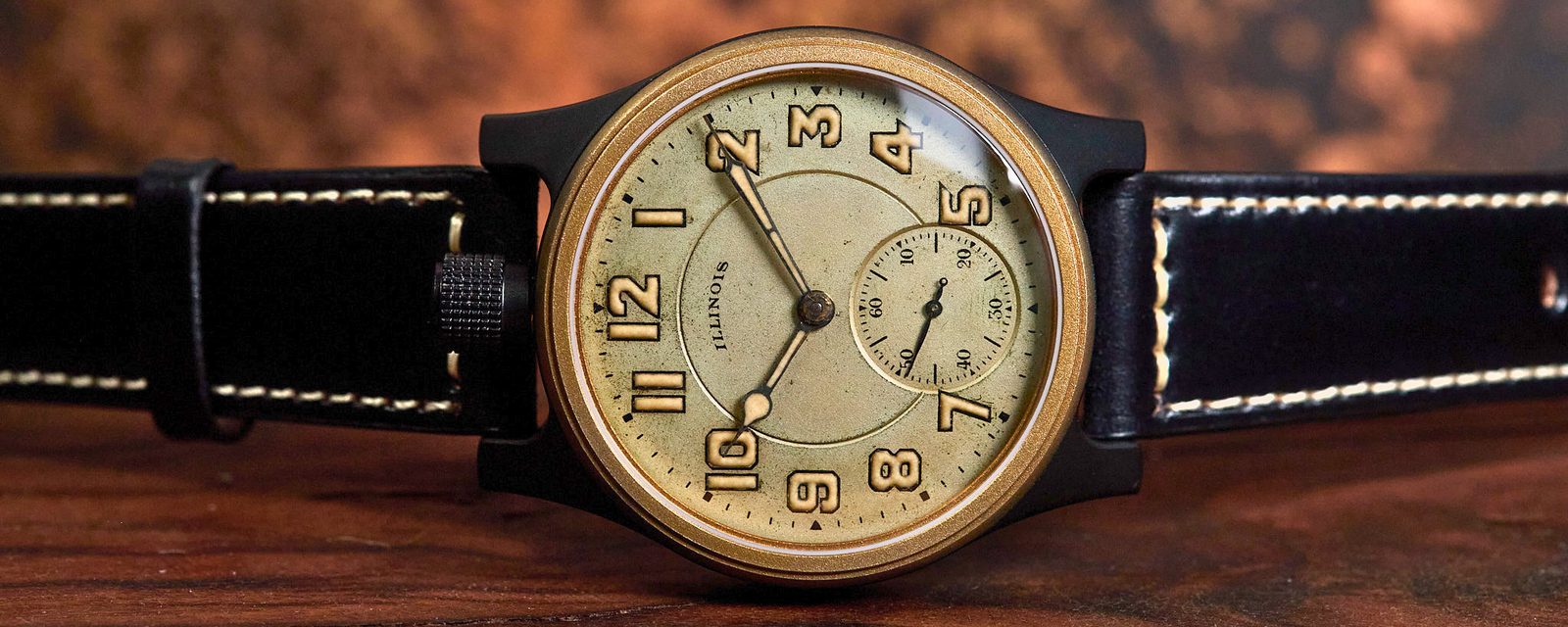A vintage-style wristwatch with a round black case, gold-toned dial, black leather strap with white stitching, and a small subdial, photographed on a wooden surface against a blurred brown background.