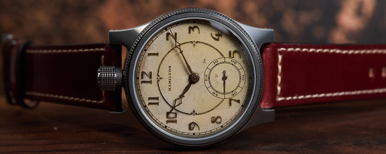 A vintage-style wristwatch with a round silver-toned case, cream dial with black Arabic numerals, and a dark red leather strap, resting on a wooden surface with a blurred rustic background.
